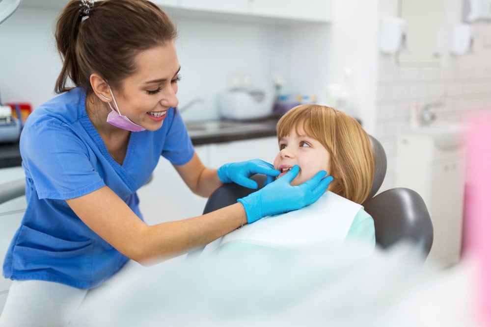 A dentist wearing blue scrubs and gloves examines the teeth of a smiling young girl sitting in a dental chair, highlighting the caring approach of Pediatric Dentistry.