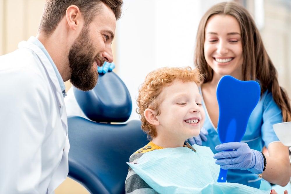A young boy at the dentist’s office smiles while looking in a blue mirror, with a male dentist and a female dental assistant beside him, highlighting the importance of preventive dentistry for maintaining healthy teeth and bright smiles. prepare your child for their first dental visit.